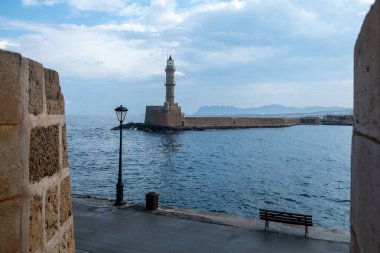 Venedik limanındaki deniz feneri, Eski Chania Girit kasabası, Yunanistan. Karanlık deniz suyu, deniz kenarında bank ve fener, bulutlu gökyüzü. Firkas Kalesinden Görünüm.