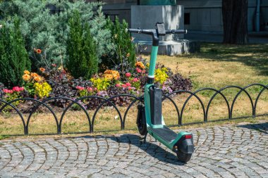 Kiralık çevre dostu elektrikli scooter şehrin çeşitli çiçek arka planlı güneşli günüyle plazanın önündeki kaldırıma park edilmiş.. 