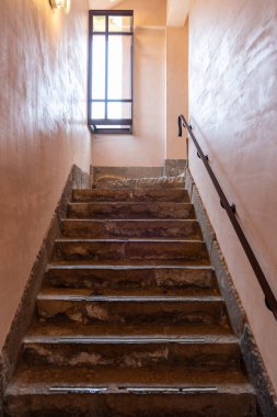 Merdivenler çıkıyor, merdivenin tepesinden ışık geliyor, duvarda metal korkuluklar var.,