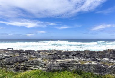 Okyanus dalgaları kayalık kıyılara çarpıyor, mavi gökyüzü dağınık bulutlarla