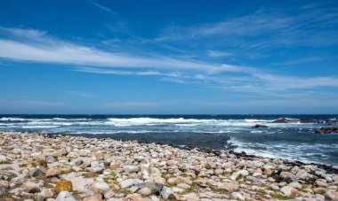 Okyanus dalgaları kayalık kıyılara çarpıyor, mavi gökyüzü dağınık bulutlarla