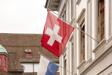 Bir binanın ön cephesinde direğe asılı İsviçre bayrağı fayanslı çatıları ve arka planında mavi-beyaz Lucerne bayrağı olan geleneksel binalar., 