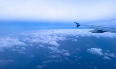 Bulutların üzerinde uçan uçak, uçağın penceresinden görünen hava manzarası, kabarık bulutlar ve mavi gökyüzü arka planı