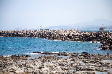 Yunanistan 'ın Girit adasındaki Rethymno limanında kayalık kıyı şeridi ve beton akıntısı. Mavi gökyüzü ve deniz, uzak dağlar, kıyı manzarası.