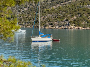 Yunanistan 'ın Ermioni kenti yakınlarında sakin bir öğleden sonra, tepe kenarı şapeli ve yemyeşil çam ağaçları olan turkuaz sularda demirlemiş huzurlu bir yelkenli..