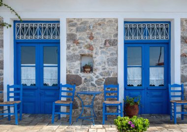 Parlak mavi ahşap kapılı Yunan taş ev cephesi, kır sandalyeleri ve çiçekli terasotta tencereleri, yaz güneşli günü