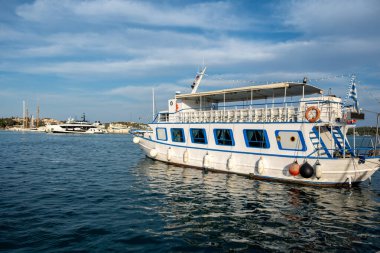 Öğleden sonra güneş ışığı altında sakin Yunan liman sularında demirlemiş beyaz ve mavi yolcu gemisi, uzak yatlar ve kıyı kenti arka planında