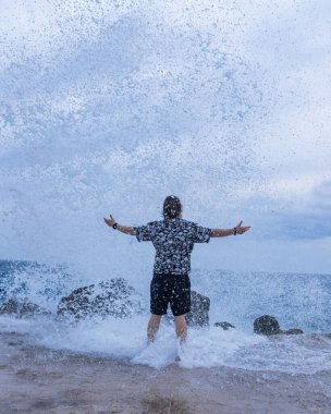 Kolları açık bir adam ıslak taş iskelede çıplak ayakla dururken güçlü okyanus dalgaları onu sarıyor. Güçlü deniz enerjisi ve bulutlu gökyüzü ile dinamik kıyı sahnesi.