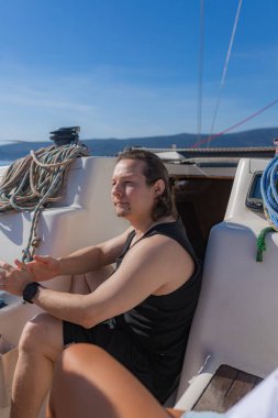 Orta boylu saçları ve akıllı saati olan genç bir adam, siyah bir atlet içinde çıplak ayakla oturuyor ve yatta şort giyiyor, vinç ipleri ve yelken ekipmanlarıyla çevrili, denize bakıyor..