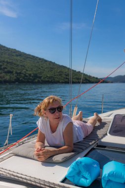 Beyaz tişörtlü ve şortlu sarışın bir kadın kameraya dönük, arka planda yeşil tepeler ve yanında mavi şişme yastıklar olan bir yatta dinleniyor..