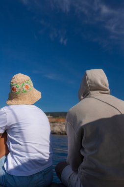 Yat güvertesinde rahat bir şekilde yan yana oturmuş, güneşli bir havanın altında sakin denize bakan iki kişinin arka görüntüsü. Yatta rahat bir şekilde oturmuş, sakin denizin tadını çıkarıyorlardı..