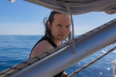 Genç adam usulca gülümsüyor. Yat donanımlarıyla çerçevelenmiş. Arka planda mavi okyanus ufku görünüyor. Nazikçe gülümseyen bir adam, siyah kolsuz tişört, yat donanımı, güneşli deniz ayarı.