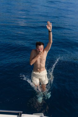 Gözlüklü gömleksiz bir erkek yattan dalarken okyanus yüzeyine çarpıyor, su dalış yaparken vücudunun her tarafına sıçrıyor..