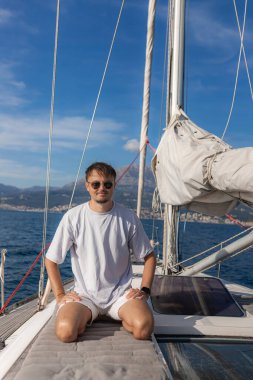 Kendine güvenen genç adam minderli tekne güvertesinde diz çöküyor, etrafı donanım ve yelkenlerle çevrili, kıyı şeridi ve arka planda dağlarla çevrili..