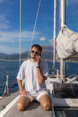 Beyaz tişört ve şort giyen şık bir adam yelkenli teknesinde telefonla konuşuyor. Sakin deniz, mavi gökyüzü, dağ kenti arka planı.