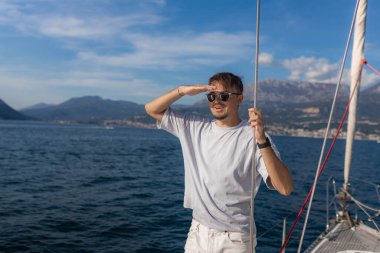 Beyaz tişört ve güneş gözlüğü takan adam, yelkenli güvertesinde elinde iple duruyor ve güneş ışığını eliyle engelliyor. Sahil şeridi ve sakin deniz manzaralı.