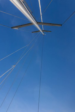 Yelken direğinin alçak açılı görüntüsü ve açık mavi gökyüzüne uzanan ipleri destekliyor. Simetrisi ve temiz çizgileri olan minimalist deniz kompozisyonu. Görünürde insan yok..