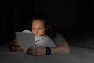 Tablet ekranı loş bir odada yüzüstü yatarken yüzünü aydınlatıyor. Bir akşam ortamında rahat ve teknoloji meraklısı bir atmosfer. Adam yatakta dinleniyor, elinde tablet tutarken ya da karanlıkta odaklanmış gözlerle kitap okuyor.