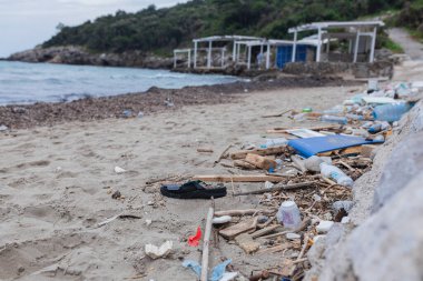 Parmak arası terlik, plastik şişeler, tahta sopalar ve okyanus kıyısındaki kumlu plajı kaplayan kumlar. Arka planda terk edilmiş yapılarla kıyı şeridi boyunca kirlilik.