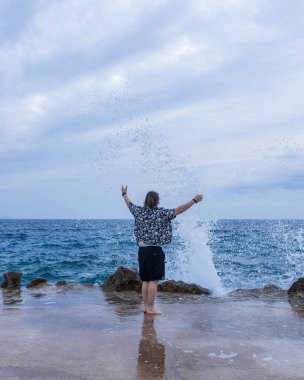 Siyah şortlu ve desenli gömlekli bir adam çıplak ayakla dev okyanus dalgalarına doğru uzanmış kayalara çarpıyor. Dramatik gökyüzü altında dinamik aksiyon sahnesi.