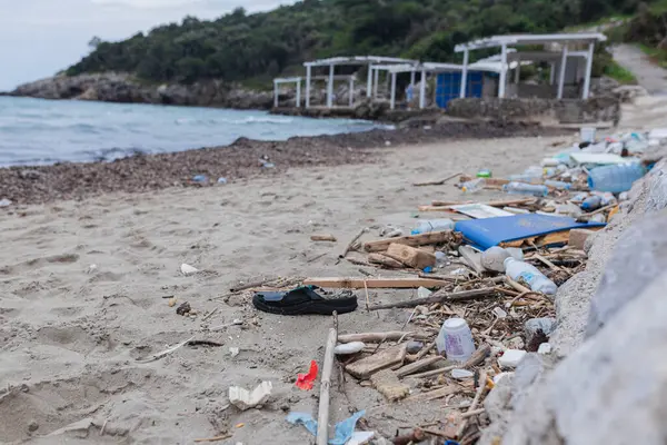 Parmak arası terlik, plastik şişeler, tahta sopalar ve okyanus kıyısındaki kumlu plajı kaplayan kumlar. Arka planda terk edilmiş yapılarla kıyı şeridi boyunca kirlilik.