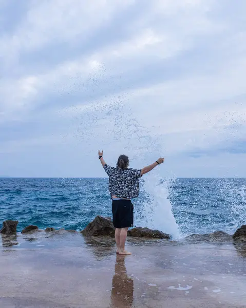 Siyah şortlu ve desenli gömlekli bir adam çıplak ayakla dev okyanus dalgalarına doğru uzanmış kayalara çarpıyor. Dramatik gökyüzü altında dinamik aksiyon sahnesi.