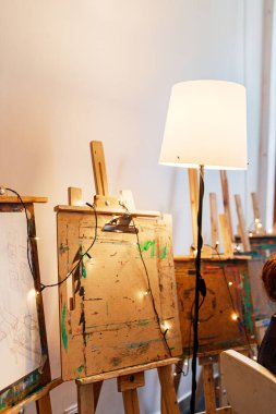 Resim sınıfındaki resim sehpalarının yakın çekimleri, sicim lambaları ve bir güvertede bir kadın resmi. Yaratıcı bir stüdyoda sanatsal bir ortam. Sanat eğitimi ve görsel hobiler için uygundur.