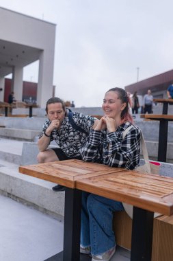 Açık şehir terasında samimi gülümseme anlarını paylaşan aynı çift. Sırt çantalı genç adam kareli tişörtlü neşeli kadına doğru eğiliyor. Temiz hatlar, taş basamaklar, yumuşak bulutlu gün ışığında modern kafeler..