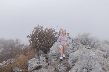Olgun bir kadın, sisli dağ ortamında büyük bir kayanın üzerinde otururken dikkatle bakıyor. Kapüşonlu ve şortlu yaşlı kadın, sis ve sonbahar çalılarıyla çevrili gri kayaların arasında oturuyor..
