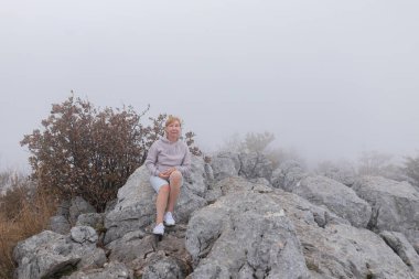 Yaşlı kadın sisli dağlık arazide huzur içinde oturuyor. Günlük giysiler giyiyor. Pastel kapüşonlu ve şortlu olgun kadın sis kaplı dağ manzarası, sakin duruş, huzur ve doğanın yalnızlığının tadını çıkarıyor..