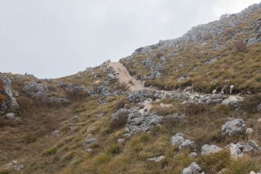 Kayalık yürüyüş yolu, sarp dağların arasından seyrek bitki örtüsüyle esiyor. Toprak patika, keskin kayaların arasından geçiyor ve bulutlu gökyüzünün altındaki çimenler soluyor. Engebeli tepe manzarası uzak yürüyüş sahneleri için ideal..