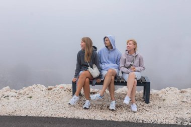 Kapüşonlu üç arkadaş sisli dağ vadilerinde yan yana oturuyorlar aynı yöne bakan bankta oturuyorlar, sis dağ vadisini kaplıyor, beyaz çakıllar ve minimalist çevre, sıcak hava koşulları için giyiniyorlar..