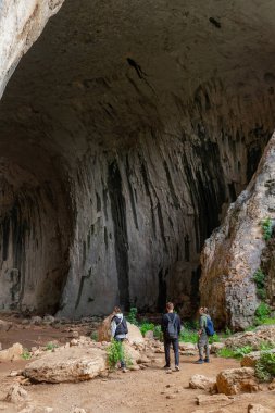 Yürüyüşçülerden oluşan bir grup dik duvar ve kaya katmanlarıyla mağara girişinde duruyor. Üç kişi Büyük Mağara 'nın tabanını keşfediyor. Dikey kaya oluşumları ve yosun çizgileri kareye hakim. Doğal dokular ve mağara jeolojisi açıkça görülüyor..
