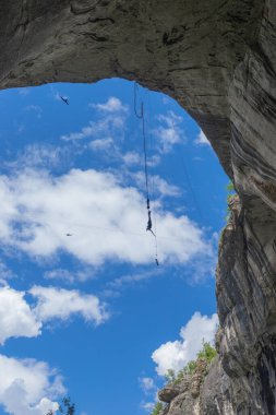 Uçsuz bucaksız mağara kemerinin ve mavi gökyüzünün altında havada asılı duran kişi. Bungee jumper havada asılı kaldı, dramatik bir gökyüzü ve kaya oluşumuyla yakalandı, aktif turizm sahnesi, özgürlük hissi.