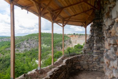 Taş duvarları ve dağ manzarası olan ahşap çatı terası. Çok farklı bir açı, aynı teras alanını gösteriyor. Ahşap çatı desteği ve ufuk boyunca uzanan panoramik yeşil arazi.