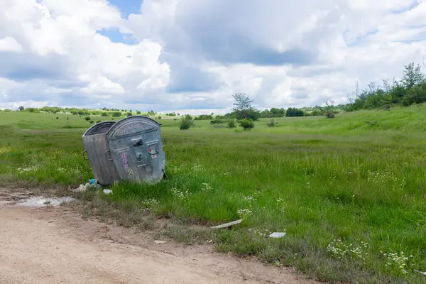 Terk edilmiş çöp tenekesi yaban çiçeği tarlasının kenarına, bulutlu gökyüzünün altına eğilmiş. Yıpranmış çöp konteyneri toprak yolun yakınına eğiliyor. Küçük beyaz papatyalar ve uzun yeşil çimenler önü bulutlu gökyüzü ile doldurur..
