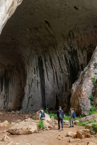 Yürüyüşçülerden oluşan bir grup dik duvar ve kaya katmanlarıyla mağara girişinde duruyor. Üç kişi Büyük Mağara 'nın tabanını keşfediyor. Dikey kaya oluşumları ve yosun çizgileri kareye hakim. Doğal dokular ve mağara jeolojisi açıkça görülüyor..