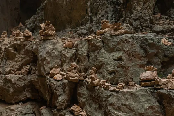 Mağaranın içinde kaya katmanları ve koyu arka kapaklı istiflenmiş taşların yatay bileşimi. Dokuma yüzeyi boyunca yatay hizada sıralanmış dengeli taşlar. Arka plandaki mağara derinlik katıyor. Keskin detay ve toprak tonları baskın gelir..