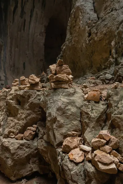 Mağaranın içinde kaya katmanları ve koyu arka kapaklı istiflenmiş taşların yatay bileşimi. Dokuma yüzeyi boyunca yatay hizada sıralanmış dengeli taşlar. Arka plandaki mağara derinlik katıyor. Keskin detay ve toprak tonları baskın gelir..
