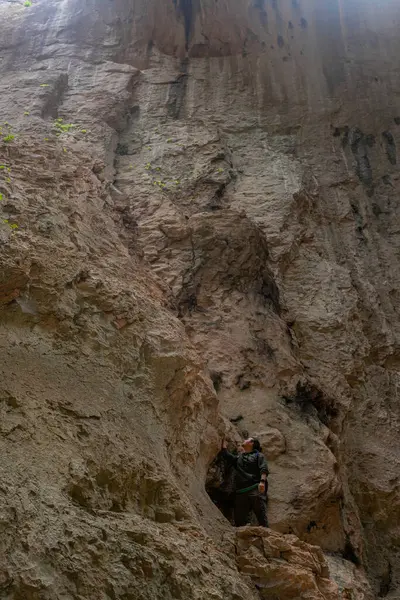 Mağaranın içinde dikey yamaç duvarında duran kişi, kaya yüzeyine doğru elini uzatıyor. Yürüyüşçü mağaranın içindeki yüksek dikey kaya yüzeyini inceliyor. Doğal duvar doku ve bitki parçalarını gösterir. Yukarıdan gelen yumuşak ışık ince bir parıltı yaratır..