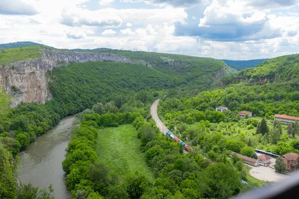 Nehir ve kireçtaşı tepesi boyunca yeşil dağ vadisinden geçen tren. Yukarıdan geniş açılı görüş, yeşil orman ve kırsal binalar tarafından çerçevelenmiş, uçurumlar ve nehir arasında seyahat eden renkli bir yük treni gösteriyor.