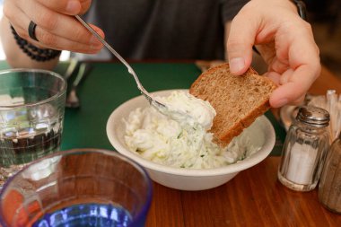 Lokanta masasında kaşık kaldırma ile kalın kremalı tzatziki 'ye batırılmış ekmek dilimi. Rustik kahverengi ekmek geleneksel salatalık yoğurt sosu, kaşık kaldırma karışımı orta havada, yeşil yer kaplaması, cam ve baharatlarla etkileşime girer..
