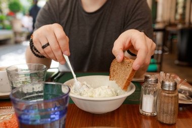 Adam açık hava restoranında çavdar ekmeğine kremalı sos ve kaşık sürüyor. Yakın plan çekimde erkek eli kaşık ve tzatziki 'nin üstünde koyu ekmek tutuyor. Doğal ışık, geleneksel ayar, çerçevede tuzluk ve biber..