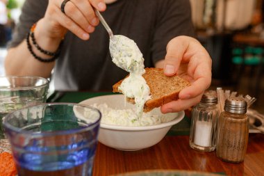 Sofrada kaşıktan koyu kremalı Yunan tzatziki ile kaplanmış Rustik kahverengi ekmek. İnsan eliyle ekmeğin üzerine salatalık parçaları serpiştirilmiş beyaz tzatziki. Tuz, biber, içme bardakları ve masa iskeleti sahnesi.