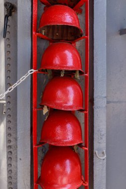 Geminin dış duvarında metal raflara istiflenmiş kırmızı kaskların yakın görüntüsü. Parlak kırmızı miğferler, gemi odasının dışındaki duvara monte edilmiş çelik raflara sıkıca yerleştirilmiş. Güvenlik önlemi için desenli yüzey ve yakınlarda zincirli erişim.
