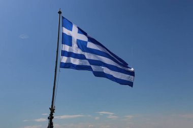 Büyük Yunan bayrağı açık mavi gökyüzünün altında deniz esintisiyle yana doğru gerilmişti. Çapraz çizgiler gökyüzünde dalgalanıyordu. Bayrak direği gemilerin kıç tarafındaki hareketlerini yakalıyor. Arkaplan gökyüzü gradyanı hafif atmosferik bulanıklık gösteriyor.