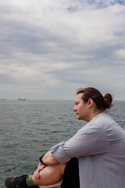 At kuyruklu bir adamın yan portresi, ahşap bir rıhtımda oturmuş okyanusu seyrediyor. Dalgın genç adamın suya oturmuş, bacak bacak üstüne atmış, açık renk tişörtlü, soluk gökyüzünün altında sakin ufuk çizgisine bakarken yakın çekimi..