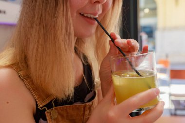 Genç kadın buzlu limon içeceğini yudumlarken gülümsüyor, görünür dişler ve günlük kafe sahnesinde bardak içiyor. Düz sarı saçlı, narenciye içen, şehir kafesinin bulanık arka planında oturan siyah pipetli, gülen bir kadın yüzü.