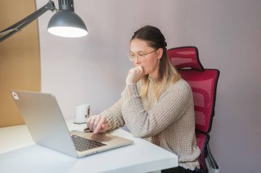 Elleri çenesinde dizüstü bilgisayara bakan bir kadın, sessiz çalışma alanında düşünceli bir ruh hali. Kırmızı ergonomik sandalyeli, sıradan kıyafetli ve evden işe giderken ekrana yansıyan yoğun bir kadın..