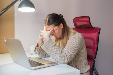 Stressed woman sitting with head bowed and hands clasped tightly.Frustrated posture at white home desk, red mesh chair and soft light, woman looking down in stress or burnout, indoor quiet scene.